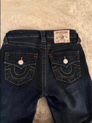 True Religion mörkblå jeans Becca - Säljer ett par mörkblå jeans från True Religion eftersom de tyvär inte har kommit till andvändning. modell Becca med mid rise bootcut. Jeansen har kontrastsömmar, dekorativa ficklock bak och silverfärgade knappar. Klassisk femficksmodell i stretchigt denimtyg. Jag har även sytt upp jeansen eftersom de var för långa, så inerbenslängden är 75cm lång💗💗