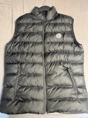 Svart dunväst från Moncler - Snygg svart dunväst från Moncler med klassisk quiltad design och dragkedja framtill. Västen har två praktiska fickor med dragkedja och Moncler-logga på bröstet. Perfekt för lager-på-lager och håller dig varm under kyliga dagar. Ett litet hål i höger ficka.