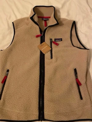 Beige fleeceväst från Patagonia - Mysig beige fleeceväst från Patagonia med hög krage och hel dragkedja framtill. Västen har svarta detaljer, tre fickor med dragkedja och röda dragflikar. Perfekt för lager-på-lager under kyliga dagar.