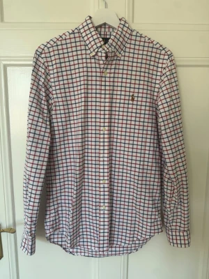 Rutig skjorta från Ralph Lauren XS - Klassisk rutig skjorta från Ralph Lauren i vitt med röda och blå rutor. Skjortan har knappar framtill, button-down krage och lång ärm. Tillverkad i mjuk bomull och har den ikoniska broderade loggan på bröstet.