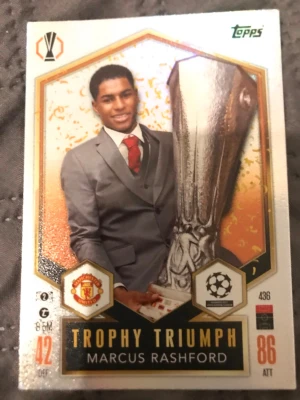 Match Attax Marcus Rashford kort - Samlarkort från Topps Match Attax med Marcus Rashford i grå kostym och röd slips, Manchester Uniteds logga och texten 'Trophy Triumph'. Kortet har guld- och svartdetaljer samt UEFA Champions League och Europa League-symboler.