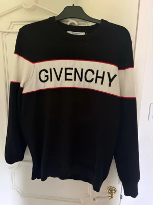 Svart stickad tröja från Givenchy - Snygg svart stickad tröja från Givenchy med vit panel och röd kant över bröstet och armarna. Stor Givenchy-logga framtill. Rund halsringning och långa ärmar. Perfekt för dig som gillar streetstyle och exklusiva märken.