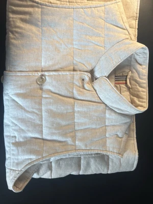 Beige quiltad linneväst med knappar - Stilren beige linneväst med quiltat mönster och ståkrage. Västen har knäppning med knappar framtill och två sidofickor. Insidan har en randig detalj vid nacken. Perfekt för lager-på-lager och enkel att matcha till olika outfits. Strlk S men passar också M.