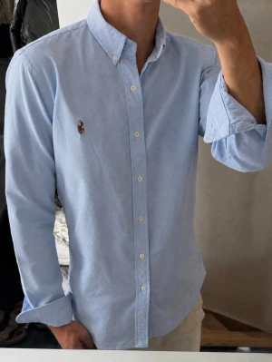 Ljusblå skjorta från Ralph Lauren - Klassisk ljusblå skjorta från Ralph Lauren med button-down krage och broderad logga på bröstet. Skjortan har långa ärmar, vita knappar och är tillverkad i bomull. Perfekt för en stilren och avslappnad look.