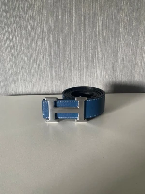 Blått skärp med H-spänne - Snyggt blått skärp med ett stort H-format metallspänne. Skärpet har vita sömmar längs kanterna som ger en stilren kontrast. Perfekt accessoar för att lyfta din outfit.