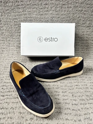 Mörkblå mocka loafers från Estro - Snygga mörkblå loafers i mocka från Estro. Skorna har en klassisk slip-on design med ljus sula och diskreta sömmar. Insidan är ljus och ger en fräsch kontrast till det mörka mockamaterialet. Perfekta för en stilren och avslappnad look.