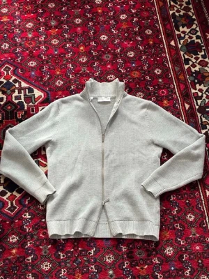Ljusgrå stickad hoodie med dragkedja - En enkel och stilren ljusgrå stickad hoodie med dragkedja framtill och huva. Tröjan har ribbade muddar vid ärmslut och nederkant, samt en mjuk och bekväm känsla. Perfekt för lager på lager under kyliga dagar.