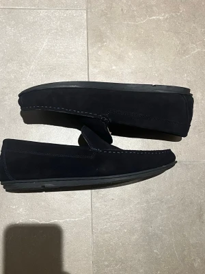 Mörkblå mocka loafers  - Säljer ett par stilrena mörkblå loafers i mocka från GANT med klassisk design och diskreta sömmar. Skorna har en låg sula i svart och är perfekta för dig som gillar en clean och tidlös look. Lätta att bara dra på sig och passar till många outfits. Loafersen är även sparsamt använda. Hör av dig vid mer frågor👊🏼🙏🏻