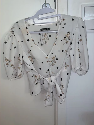Vit blus med knyt och prickar  - Supersöt vit blus från Boohoo med svarta prickar och blommigt mönster. V-ringad omlottmodell med knyt i midjan och puffiga korta ärmar. Perfekt för dig som gillar romantisk och trendig stil.