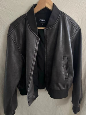 Svart skinnjacka från ONLY - Snygg svart skinnjacka från ONLY med klassisk bombermodell. Jackan har ribbade muddar vid ärmslut och midja, dragkedja framtill och två fickor. Perfekt för en cool och stilren look.