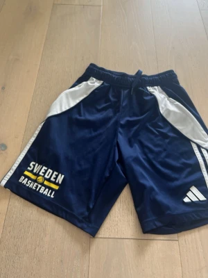 Blå Sweden Basketball shorts Adidas - Snygga basketshorts i mörkblått och vitt från Adidas med Sweden Basketball-tryck och dragkedjeförsedda fickor. Storlek xs 