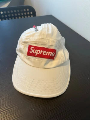 Vit Supreme keps med röd logga - Säljer en vit keps från Supreme med den klassiska röda boxloggan framtill. Kepsen har böjd skärm, justerbar rem bak och meshpaneler på sidorna för extra ventilation. Perfekt för dig som vill ha en streetwear-look.