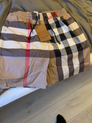 Bb shorts - Säljer ett par rutiga shorts med snörning i midjan. Shortsen har ett klassiskt beige, svart, vitt och rött rutmönster och elastisk midja för skön passform. Perfekta för sommaren och riktigt snygga till en enkel t-shirt. Bb shorts storlek L passar M
