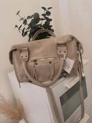 Beige handväska med gulddetaljer - Snygg beige handväska i mockaimitation med guldfärgade metalldetaljer och dragkedja framtill. Väskan har både handtag och en lång axelrem, samt dekorativa spännen och nitar. Perfekt för dig som vill ha en stilren och trendig accessoar.