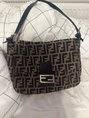 Fendi Baguette monogram handväska - Säljer en ikonisk Fendi Baguette handväska i brunt och svart med klassiskt FF-monogram över hela väskan. Väskan har ett justerbart axelband i svart läder och en silverfärgad spänne framtill med Fendi-logga. Materialet är canvas med detaljer i läder.