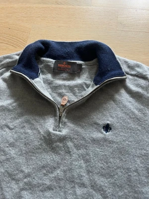 Grå halvzip tröja från Morris - Snygg grå tröja från Morris med halv dragkedja och mörkblå krage. Tröjan har ett diskret blått broderat emblem på bröstet och ribbade muddar vid ärmslut och nederkant. Perfekt för en stilren och avslappnad look. Även ett litet hål på baksidan därav skicket