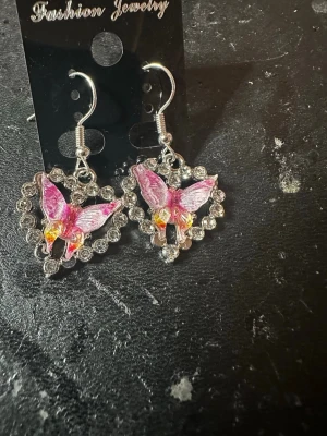 Hjärtfomade örhängen med fjärilar - Säljer ett par söta örhängen med hjärtformad ram dekorerad med glittrande stenar och en rosa fjäril i mitten. Örhängena har krokar i silverfärg och detaljer i rosa och gult. Perfekta för dig som gillar unika accessoarer med lite extra bling.