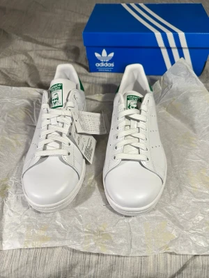 Adidas Stan Smith vita sneakers - Klassiska vita Adidas Stan Smith sneakers med gröna detaljer på hälen och tungan. Skorna har en stilren design med perforerade ränder på sidorna och är tillverkade i slätt läder. Perfekta för en clean och tidlös look.