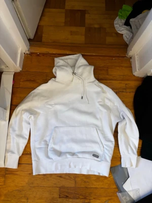Vit hoodie med magficka och huva - Snygg vit hoodie med stor magficka och justerbar huva med dragsko. Klassisk och enkel design, perfekt för en avslappnad stil. Tillverkad i mjukt bomullstyg för extra komfort. Liten patchdetalj nedtill på fickan.