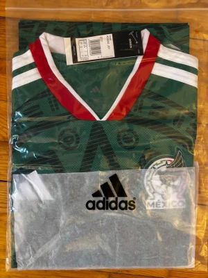 Grön Mexiko fotbollströja Adidas - Säljer en grön Mexiko fotbollströja från Adidas med vit och röd V-ringning. Tröjan har tryckt Mexiko-emblem på bröstet och klassiska Adidas-detaljer. Perfekt för dig som älskar fotboll och vill stötta Mexikos landslag.