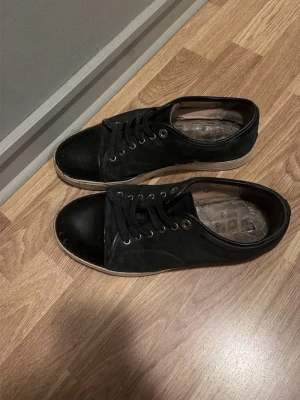 Svarta låga sneakers med gummitå - Säljer ett par svarta sneakers med klassisk låg profil och rundad tå i gummi. Skorna har svarta snören, metallöljetter och vit sula. Materialet är en mix av tyg och gummi vilket ger en clean och enkel look. 