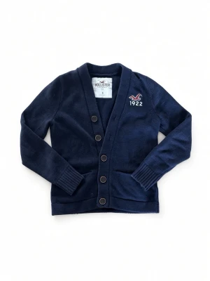 Mörkblå kofta från Hollister - Mörkblå kofta från Hollister med broderad logga och årtal 1922 på bröstet. Koftan har v-ringning, ribbade muddar och knäppning med fyra knappar framtill. Perfekt för lager-på-lager och en chill stil. Bredd: 36cm, Längd: 60cm.