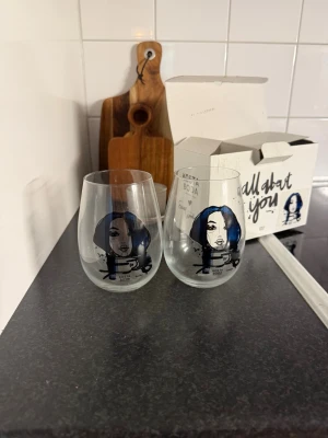 Kosta Boda glas med porträttmotiv - Två glas från Kosta Boda med unikt porträttmotiv i blått och svart. Glasen har en modern, rundad form och är dekorerade med illustration av ett ansikte. Perfekta för dryck eller som dekoration. Originalförpackning medföljer.