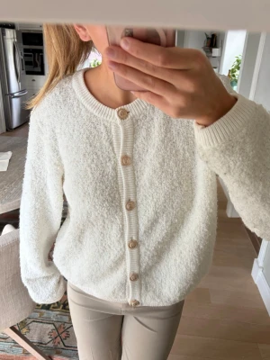 Vit fluffig kofta - Mysig vit kofta med fluffig textur och rund halsringning. Koftan har långa ärmar, ribbade muddar och knäpps framtill med snygga guldiga knappar. 