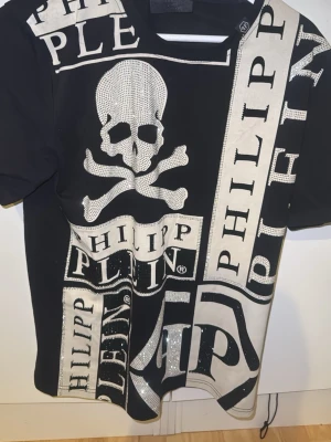 Svart t-shirt med döskalle och logga - Snygg svart t-shirt med stora vita tryck, döskalle och PHILIPP PLEIN-logga över hela plagget. T-shirten har korta ärmar och glittriga detaljer på framsidan som ger extra edge. Perfekt för dig som gillar statement-plagg och streetstyle.