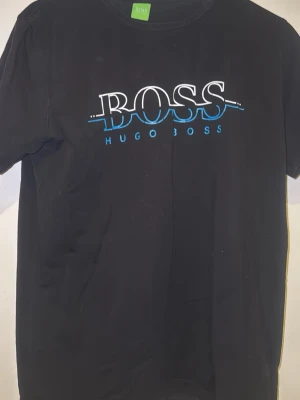 Svart t-shirt från Hugo Boss - Snygg svart t-shirt från Hugo Boss med stort vitt och blått BOSS-tryck på bröstet. Klassisk rund hals och korta ärmar. Perfekt för dig som gillar stilrena och trendiga plagg med tydlig logga.