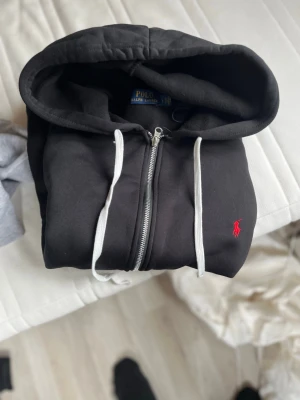 Svart hoodie från Polo Ralph Lauren - Svart hoodie med dragkedja från Polo Ralph Lauren. Klassisk design med vit dragsko i huvan och röd broderad logga på bröstet. Mjuk insida och praktisk huva gör den perfekt för en chill och stilren look.är i L men passsr m