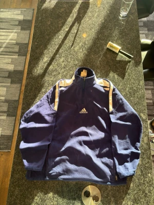 Blå fleece-tröja från Adidas - Mysig blå fleece-tröja från Adidas med vita ränder på axlarna och halv dragkedja framtill. Perfekt för kyliga dagar och har en sportig vibe. Adidas-logga broderad på bröstet och hög krage för extra värme.