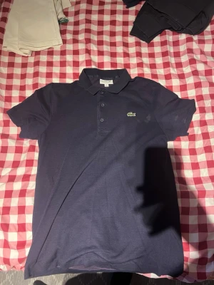  Lacoste piké  - Snygg mörkblå pikétröja från Lacoste med klassisk krage och knappar framtill. Tröjan har några fläckar som går bort i tvätten enkelt. Pris kan diskuteras!
