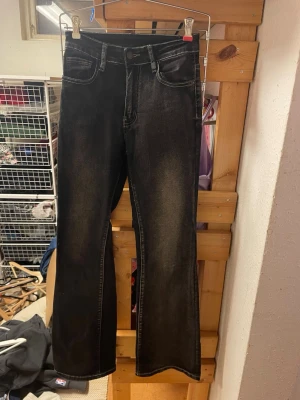 Svarta bootcut jeans med broderade fickor - Säljer ett par svarta bootcut jeans med snyggt slitna detaljer och vita kontrastsömmar. Baksidan har två fickor med dekorativa broderier och knappar. Jeansen har klassisk femficksmodell och stängs med knapp och dragkedja. Perfekta för en trendig och avslappnad look.