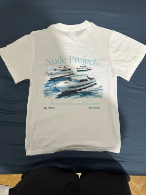 Vit Nude Project t-shirt med tryck - Vit t-shirt från Nude Project med tryck av båtar och texten 'Never trust a millionaire' på både fram- och baksida. Klassisk passform och rund hals. Perfekt för dig som gillar streetwear och unika prints.