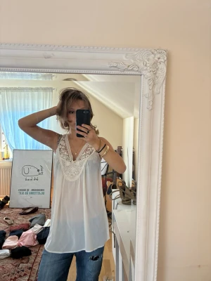 Vit spetsdetaljerad linnetopp - Säljer en luftig vit linnetopp med breda axelband och v-ringning. Toppen har vackra virkade spetsdetaljer runt halsen och är tillverkad i ett tunt, lätt material som passar perfekt till jeans eller kjol.