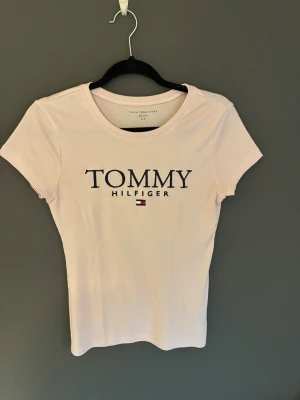 Ljus rosa T-shirt från Tommy Hilfiger - Snygg ljus rosa T-shirt från Tommy Hilfiger med klassisk logga och liten flagga framtill. T-shirten har rund halsringning och korta ärmar. Tillverkad i mjuk bomull för en bekväm känsla och sitter snyggt med en normal passform.