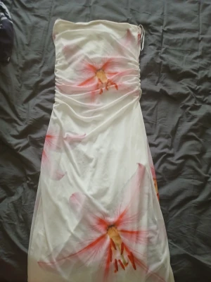 Vit långklänning med blommor Gina Tricot - Säljer en vit axelbandslös långklänning från Gina Tricot med stora blommiga mönster i rosa och orange toner. Klänningen har en figurnära passform upptill och faller snyggt nedåt. Perfekt för sommarens alla tillfällen!