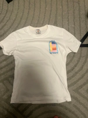 Vit Casa Blanca t-shirt med tryck - Säljer en vit t-shirt från Casa Blanca med ett färgglatt grafiskt tryck på bröstet. T-shirten har rund hals och korta ärmar. Perfekt för dig som gillar stilrena plagg med en twist.