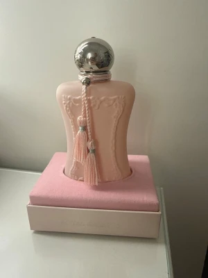 Delina parfym från Parfums de Marly - Lyxig parfymflaska i matt ljusrosa med silverfärgat klotlock och dekorativa tofsar. Kommer i elegant rosa ask med reliefdetaljer och logotyp. Doften Delina är känd för sin feminina och exklusiva känsla. Köpte den från Vinted därav billiga priset. Sprayat fåtal gånger 