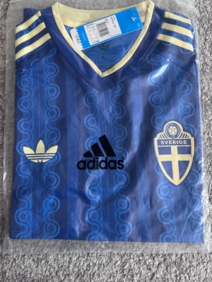 Blå Sverige tröja från Adidas - Säljer en blå Sverige tröja från Adidas med retroinspirerat mönster, gula detaljer och broderad landslagssköld. Tröjan har korta ärmar, V-ringning och tre gula ränder på axlarna. Perfekt för dig som vill stötta Sverige med stil.