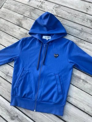 Blå hoodie från comme des garcon💙 - Säljer en blå hoodie med dragkedja och huva. På bröstet sitter en svart hjärtformad patch med ögon, vilket ger en unik och cool look. Hoodien har två fickor framtill och ribbade muddar vid ärmslut och nederkant. Perfekt för dig som gillar streetwear.