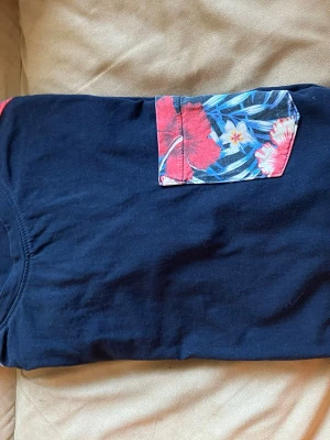 Svart t-shirt med blommig ficka - Svart t-shirt med en färgglad bröstficka i blommigt mönster i rött, blått och vitt. T-shirten har klassisk passform och är tillverkad i mjuk bomull, perfekt för en avslappnad och stilren look.