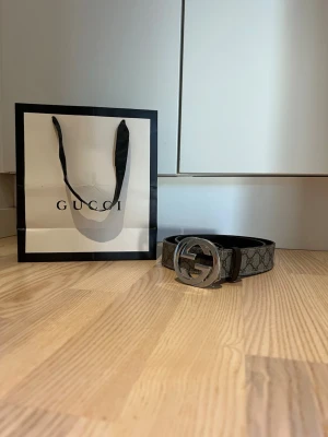 Gucci bälte med GG-spänne - Snyggt Gucci bälte med klassiskt GG-spänne i silver och monogrammönstrad rem i grått och svart. Perfekt accessoar för att lyfta vilken outfit som helst. Kommer med originalpåse. Bältet är äkta men det billiga priset beror på att jag har gjort ett eget hål med en håltagare i bältet för att det ska passa mig och jag har dessvärre inte kvar kvittot eller box. 