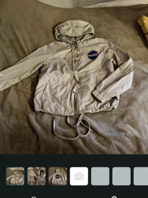 Beige vindjacka med NASA-tryck - Snygg beige vindjacka från Divided med NASA-logga på bröstet. Jackan har huva, dragsko i midjan och muddar vid ärmsluten. Tillverkad i lätt material som passar perfekt för blåsiga dagar. Enkel och stilren design med dragkedja framtill.