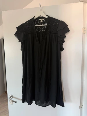 Svart kort klänning med volangärm - Svart kort klänning från H&M med v-ringning, broderade detaljer upptill och volangärmar. Klänningen är luftig och har en avslappnad passform, perfekt för varma dagar. Tillverkad i ett lätt material som känns skönt mot huden.