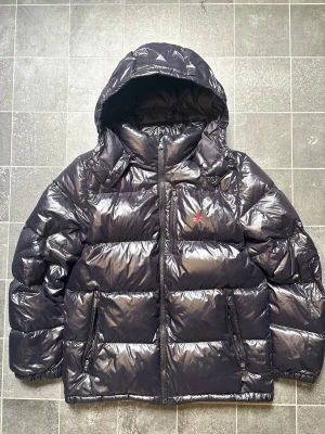Svart pufferjacka med huva från Polo Ralph Lauren - Säljer en svart, glansig pufferjacka från Polo Ralph Lauren med huva och dragkedja framtill. Jackan har två fickor med dragkedja och den klassiska röda Polo-loggan broderad på bröstet. Perfekt för kalla vinterdagar och riktigt skön att bära.