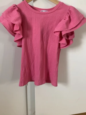 Rosa ribbad topp med volangärm Zara - Supersöt rosa topp från Zara med ribbad struktur och stora volangärmar som ger en extra touch. Perfekt för dig som vill sticka ut lite extra! Rund halsringning och mjukt material som känns skönt mot huden.