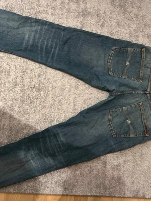 Blå raka jeans med slitningar - Säljer ett par blå jeans med raka ben och tydliga slitningar framtill och baktill. Jeansen har klassiska fem fickor, kontrastsömmar och en något tvättad look. Materialet är denim i bomull. Perfekta för en avslappnad och trendig stil.