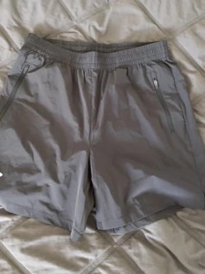 Grå träningsshorts med dragkedja - Snygga grå träningsshorts med elastisk midja och snörning. Shortsen har två fickor med dragkedja och är tillverkade i ett lätt syntetmaterial som andas. Perfekta för aktiva dagar eller gymmet.
